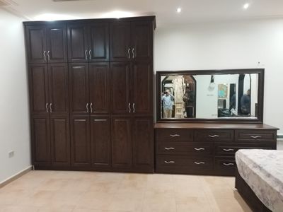 A premium full bedroom furniture set featuring a rich natural wood oak finish, including a clean-lined bedframe with an integrated headboard, two matching minimalist nightstands, a spacious multi-drawer dresser, and a large matching wardrobe closet.