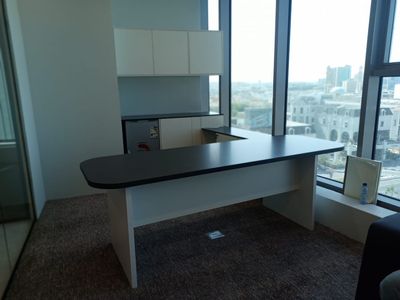 A matching office furniture suite including an executive main desk, a side return table with drawers, and a background storage counter with wall-mounted cupboards.