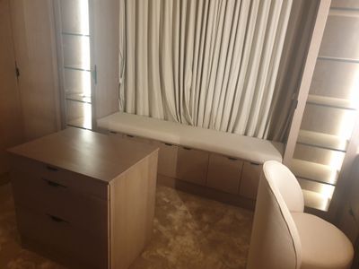 A luxury walk-in closet furniture suite featuring tall twin shelving units and an integrated window bench seat with lower cupboards, a matching wardrobe closet, a central storage island table with drawers, and a coordinating dresser chair.